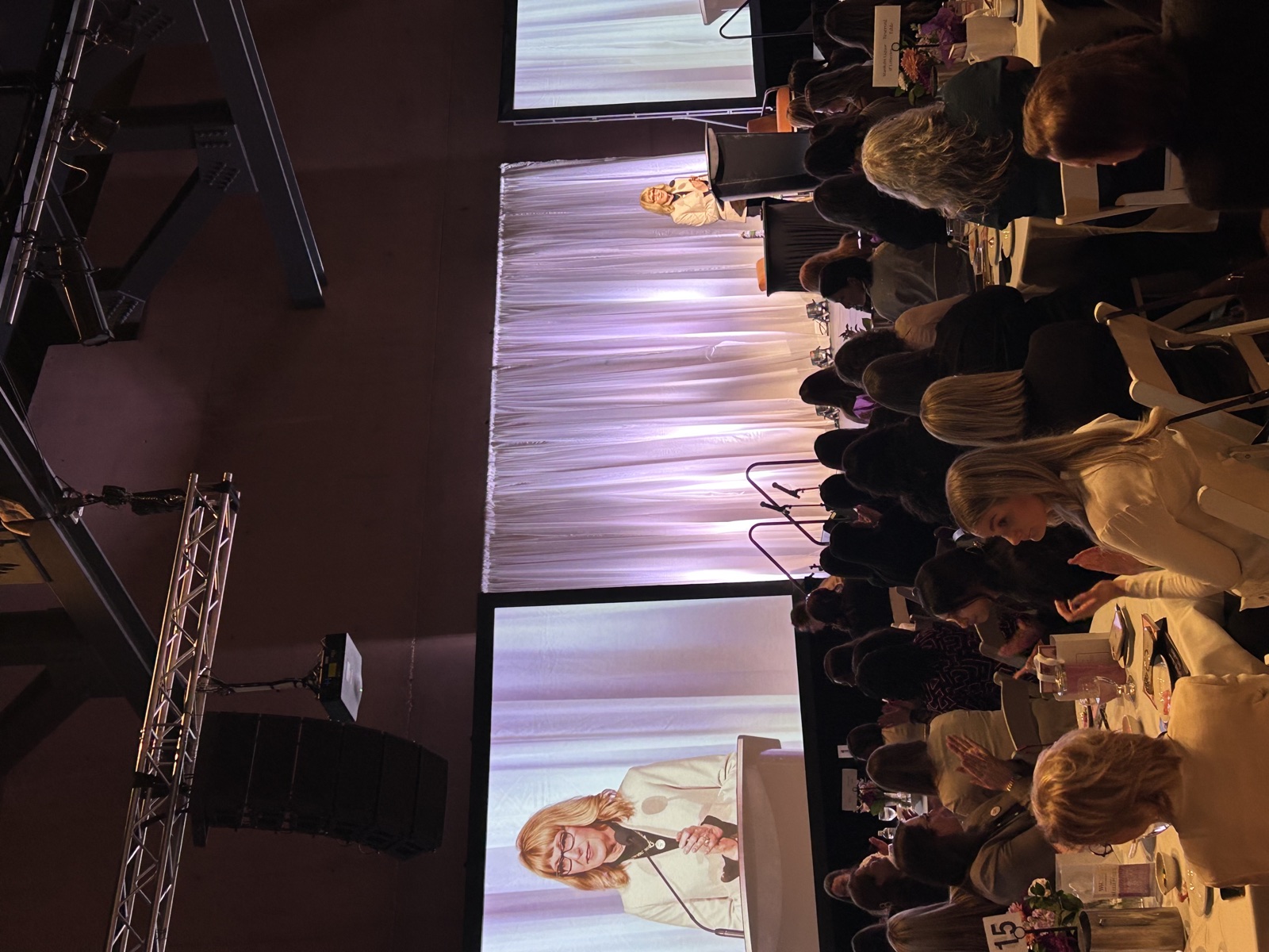 An audience seated at round tables watching Esther Findlay speak.